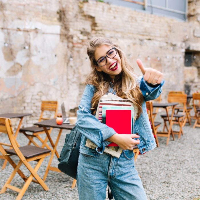 Stylish hipster girl in the retro jeans suit posing in front of the old brick wall. Trendy young woman with bag standing next to old building. Street fashion concept, wearing glasses
